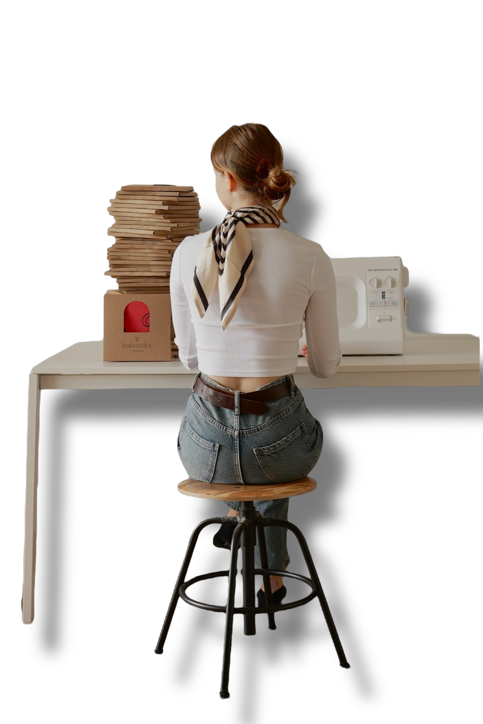Kobieta siedząca tyłem przy stole przed maszyną do szycia z zapakowanymi apaszkami po prawej stronie